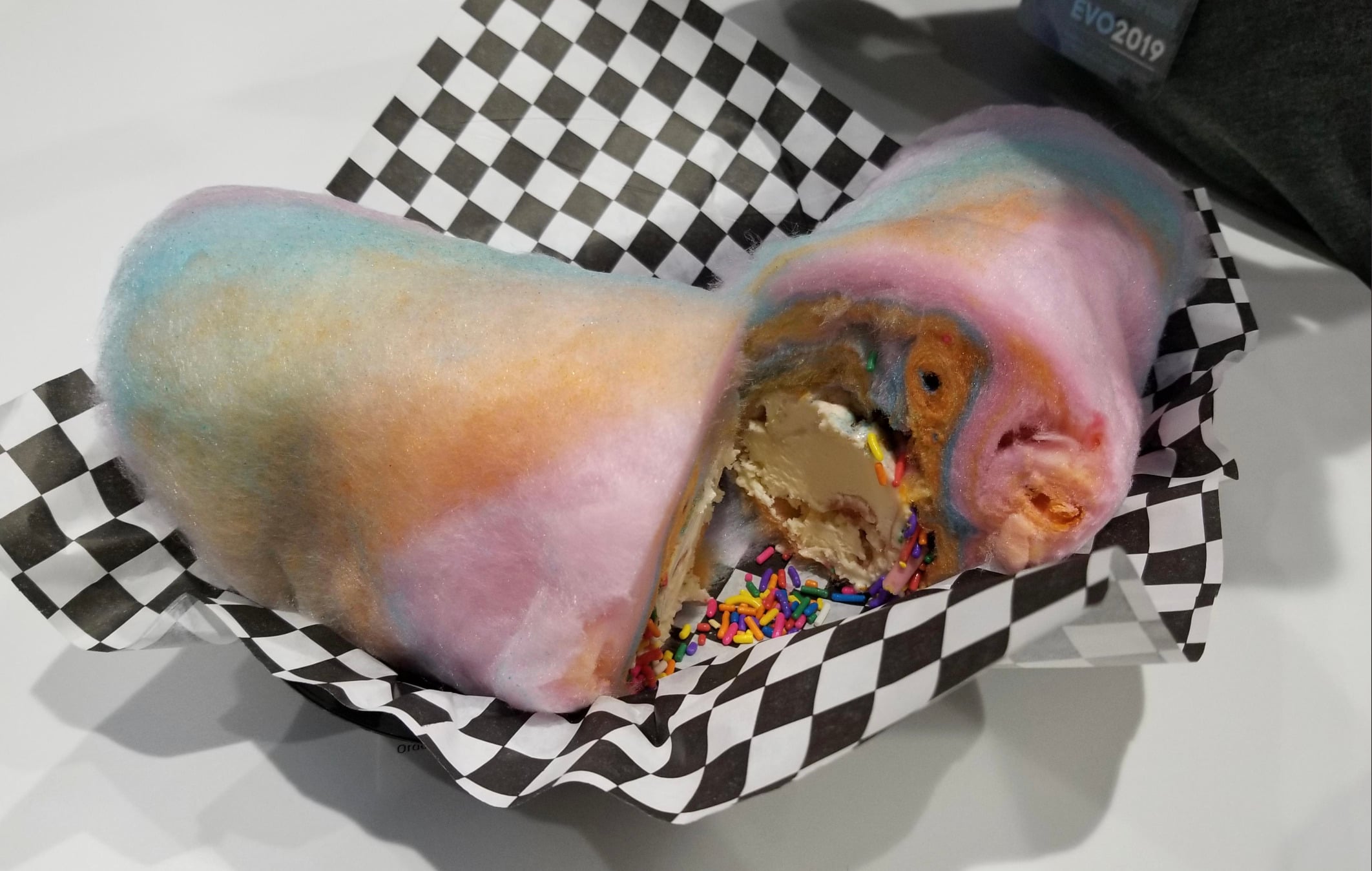 A dessert wrapped in colorful cotton candy, filled with ice cream and rainbow sprinkles, served in a black and white checkered paper-lined basket.