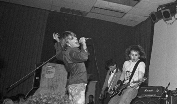 Black and white photo of a band performing on stage; the vocalist sings passionately into a microphone while two guitarists play, one in a suit jacket. Stage lights and an amplifier are visible in the background.