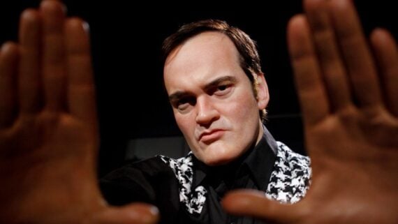 A man with dark hair and a serious expression holds his hands up, framing his face as if looking through a camera viewfinder. He wears a patterned black and white jacket over a black shirt against a dark background.