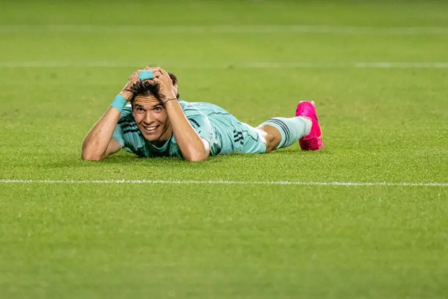 A soccer player in a light blue uniform and pink shoes lies face down on the grass field, holding his head with both hands and smiling, possibly expressing relief or disbelief.