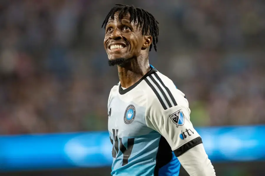 A soccer player in a blue and white jersey with black sleeves smiles while looking upward during a match. The background is blurred with stadium lights and spectators visible.