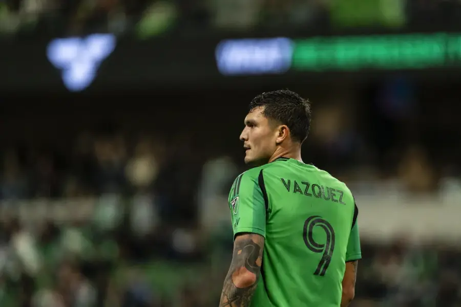 A soccer player wearing a green jersey with the name "Vazquez" and the number 9 on the back stands on the field, looking to the side with a stadium crowd blurred in the background.