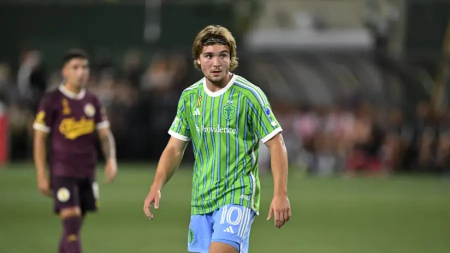 A soccer player in a green and blue striped jersey with the number 10 stands on the field during a match, with another player in a maroon uniform blurred in the background.