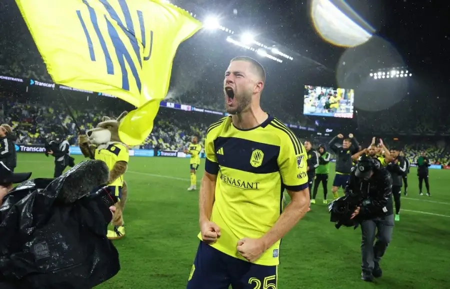 A soccer player in a yellow and navy uniform celebrates energetically on the field, clenching his fists and shouting, with a large yellow flag waving and cameras capturing the moment. Fans and teammates are visible in the background.