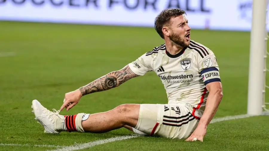 A soccer player in a white FC Dallas uniform sits on the field holding his leg, appearing in pain. He wears a captain's armband and has visible tattoos on his arm. The background shows part of an advertisement board.