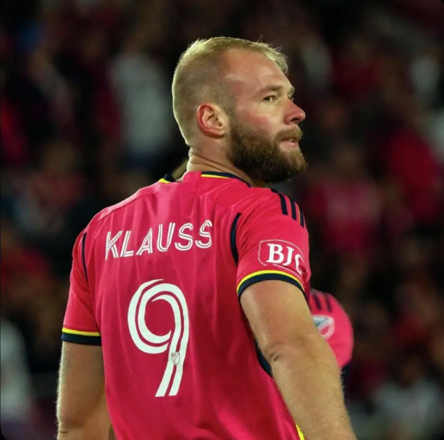 A soccer player with a blond beard wearing a red jersey with the name "KLAUSS" and the number 9 looks over his shoulder during a game. The background is blurred with spectators.
