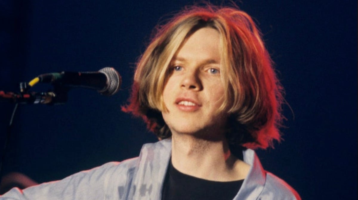 A young person with shoulder-length blond hair stands in front of a microphone, wearing a light blue shirt over a black top, under stage lighting with a dark background.