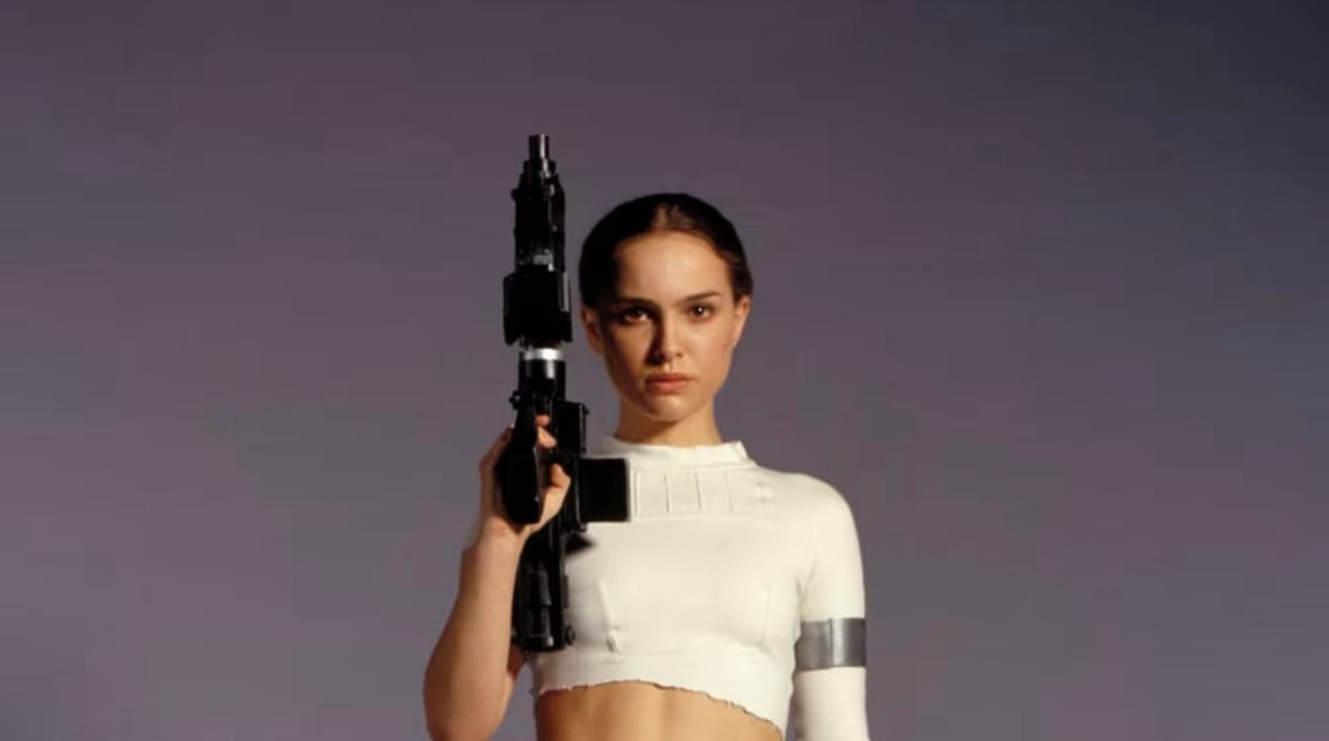 A woman in a white cropped top holds a large black sci-fi blaster upright beside her face, standing against a plain background and looking forward with a serious expression.