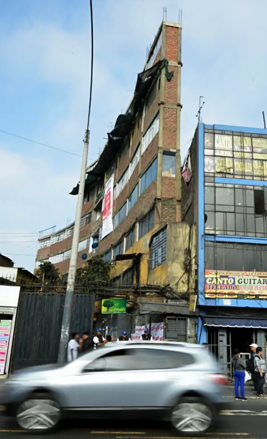 A tall, narrow, unfinished building with exposed brick and several signs stands on a busy street. A gray car passes in the foreground, and blurred pedestrians walk along the sidewalk beneath a cloudy sky.