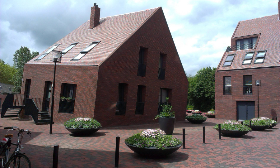 Two modern brick buildings with steeply pitched roofs and large windows, surrounded by round planters filled with flowers, on a paved courtyard. A bicycle is parked on the left. Trees and cloudy sky in the background.