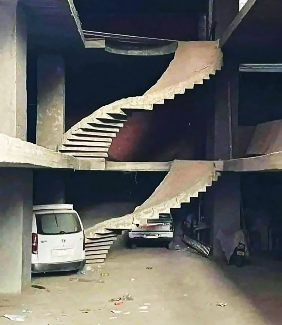 A pair of unfinished, floating concrete staircases in a parking area, not connecting to any floor, with two cars parked below and construction debris scattered on the ground.
