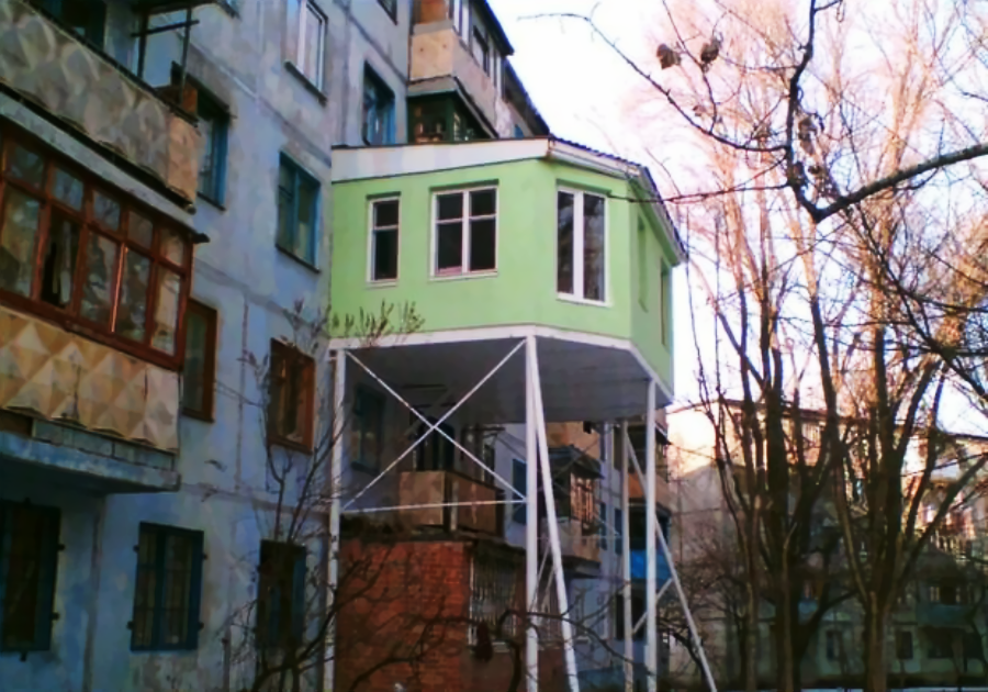 A green, box-shaped room is oddly attached to the side of an old apartment building, elevated on thin white stilts above the ground and lower balconies. Leafless trees surround the structure.