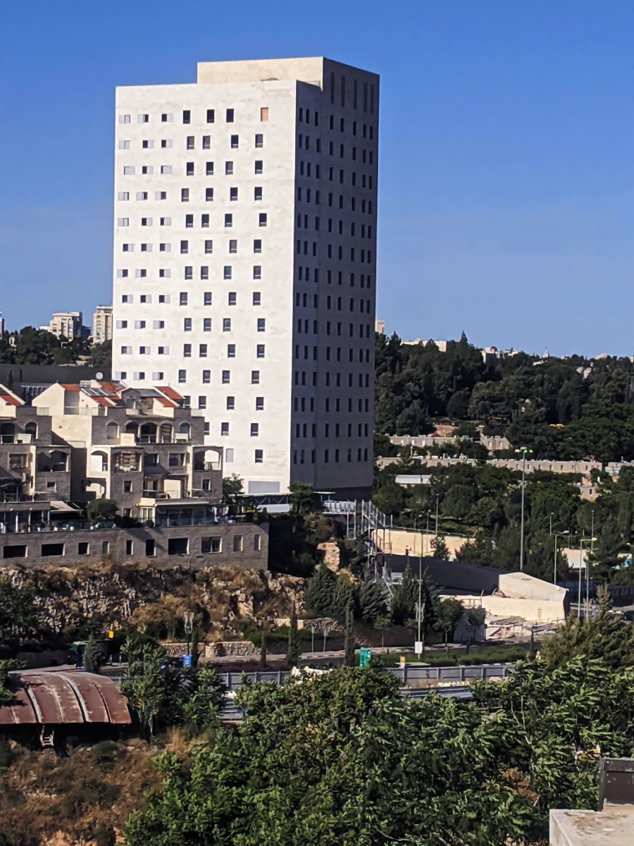 A tall, rectangular, white high-rise building stands behind shorter residential buildings, surrounded by greenery under a clear blue sky. A road and trees are visible in the foreground.