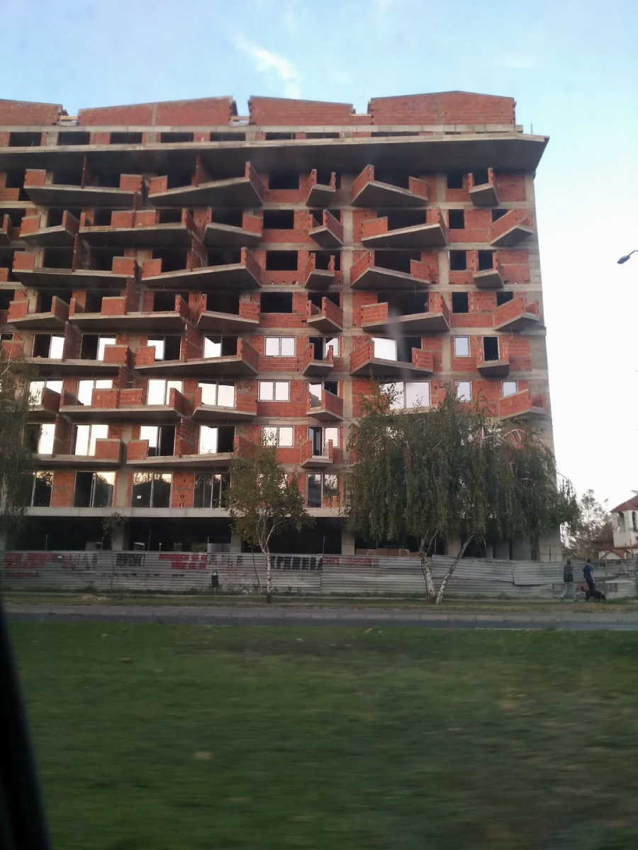 A partially constructed building with irregular and misaligned windows and balconies, creating a chaotic and uneven appearance. There are trees and grass in the foreground, and a few people walking by.