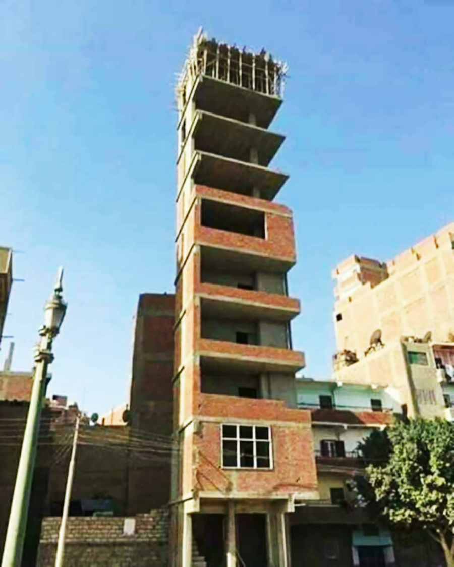 A tall, narrow, unfinished brick building with several floors but no windows or doors, standing between other structures under a clear blue sky.