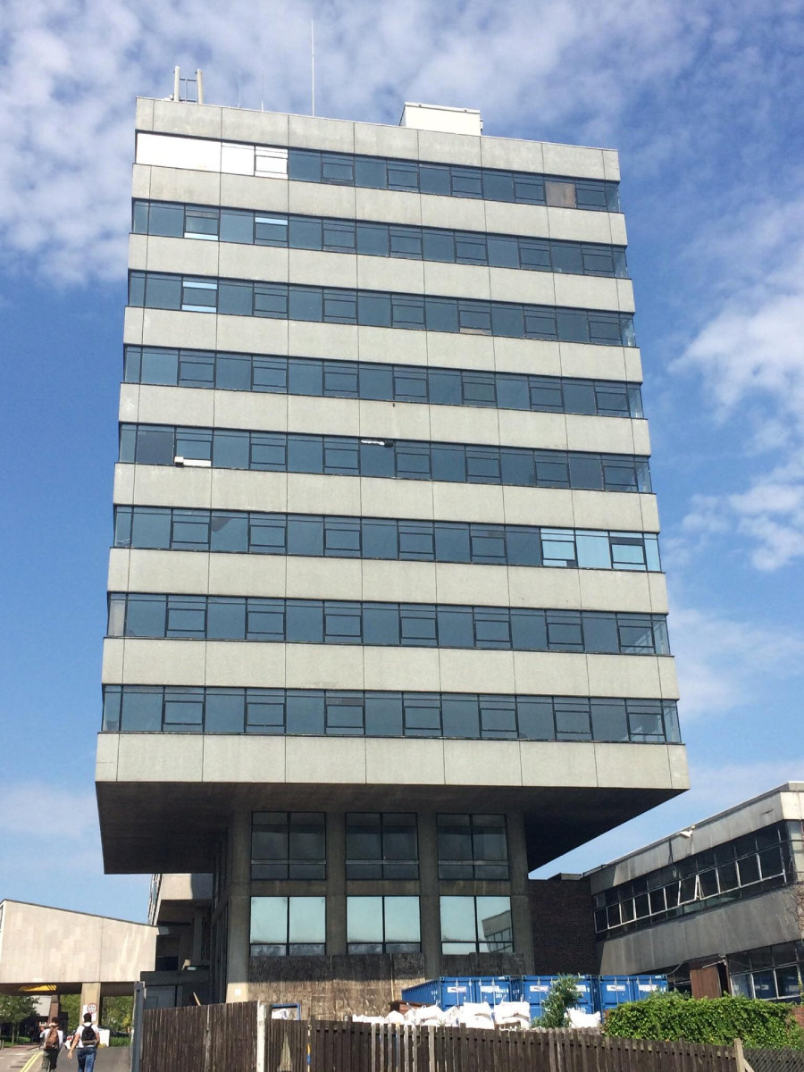 A tall, modern office building with many windows and an overhanging upper structure stands against a partly cloudy sky, with people walking and adjacent low buildings visible at its base.