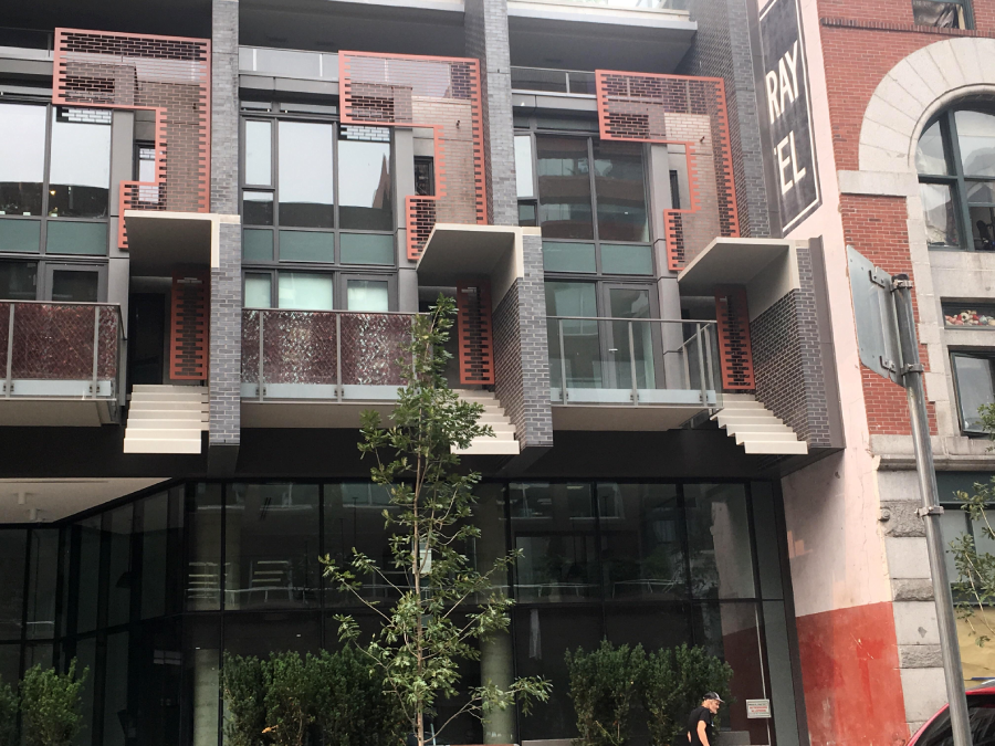 A modern building facade with large glass windows, geometric red metal accents, and protruding balcony structures. There are plants in front and a person walking on the sidewalk.
