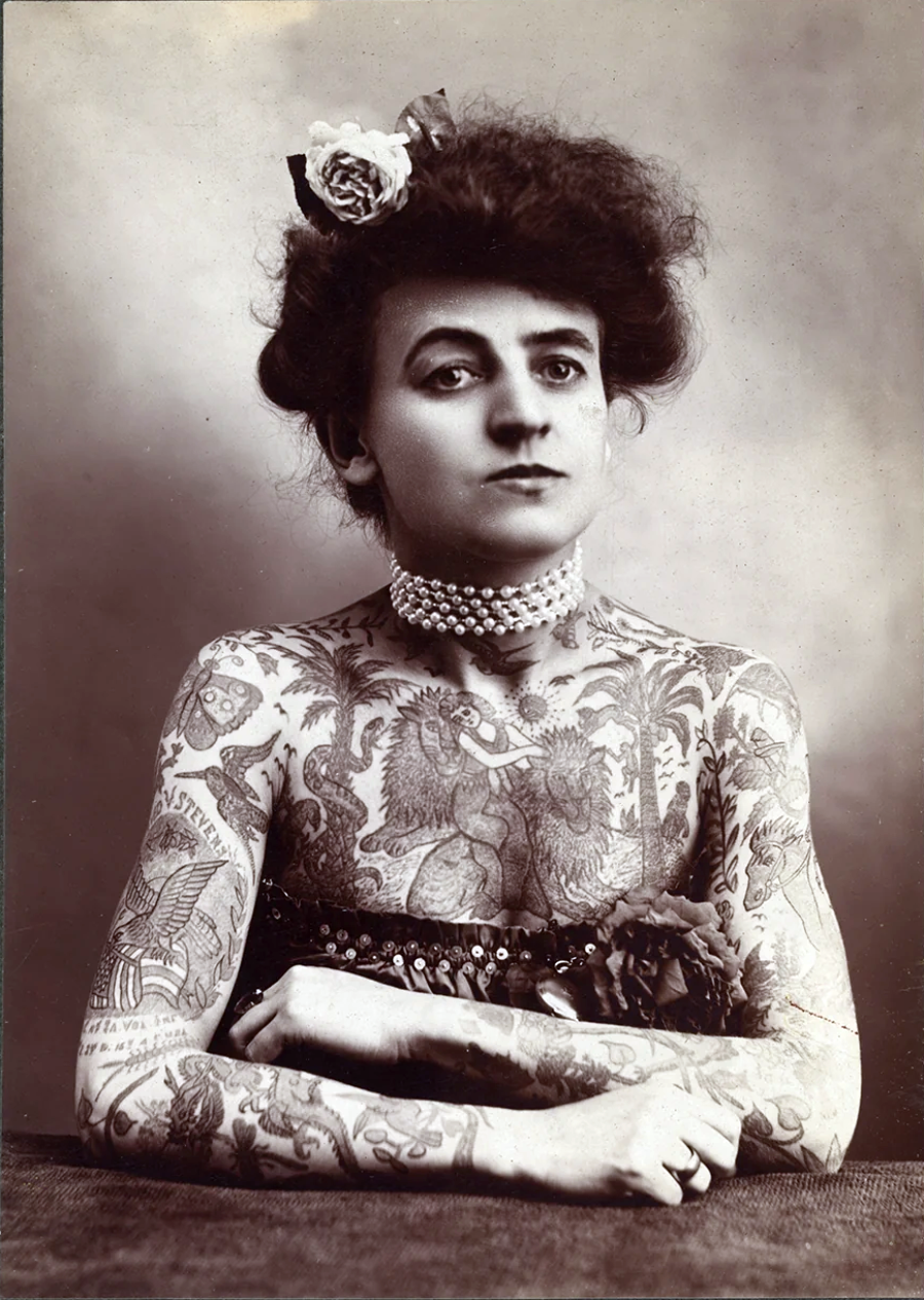 A black-and-white vintage portrait of a woman with her arms crossed, wearing a pearl necklace and flower in her hair. Her upper body and arms are covered in intricate tattoos.