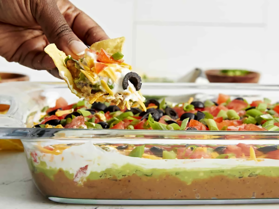 A hand scoops a tortilla chip into a glass dish filled with layered dip, including beans, guacamole, sour cream, tomatoes, cheese, green onions, and sliced black olives.