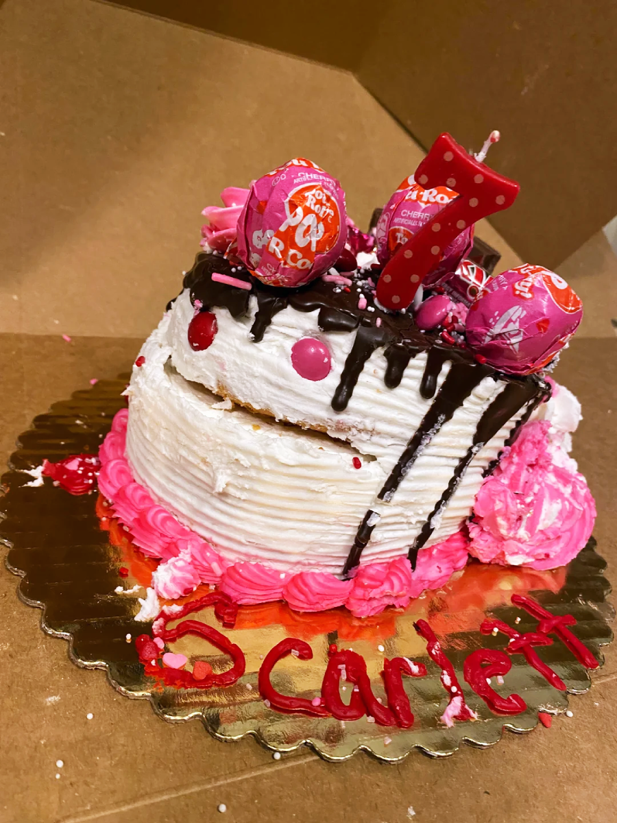 A white frosted birthday cake with pink decorations, Tootsie Pops on top, a pink number 7 candle, and “Scarlett” written in red icing on a gold tray. The cake is slightly tilted inside a cardboard box.
