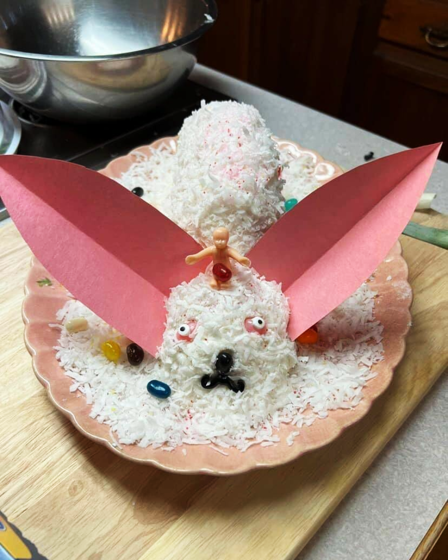 A bunny-shaped cake covered in white coconut flakes, with large pink paper ears, jellybean decorations, and a small doll figure on its head, displayed on a plate.