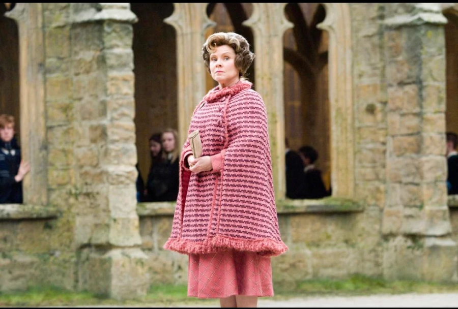 A woman with curled hair, wearing a pink patterned cape and holding a book, stands in a stone courtyard with arched windows and people in the background.