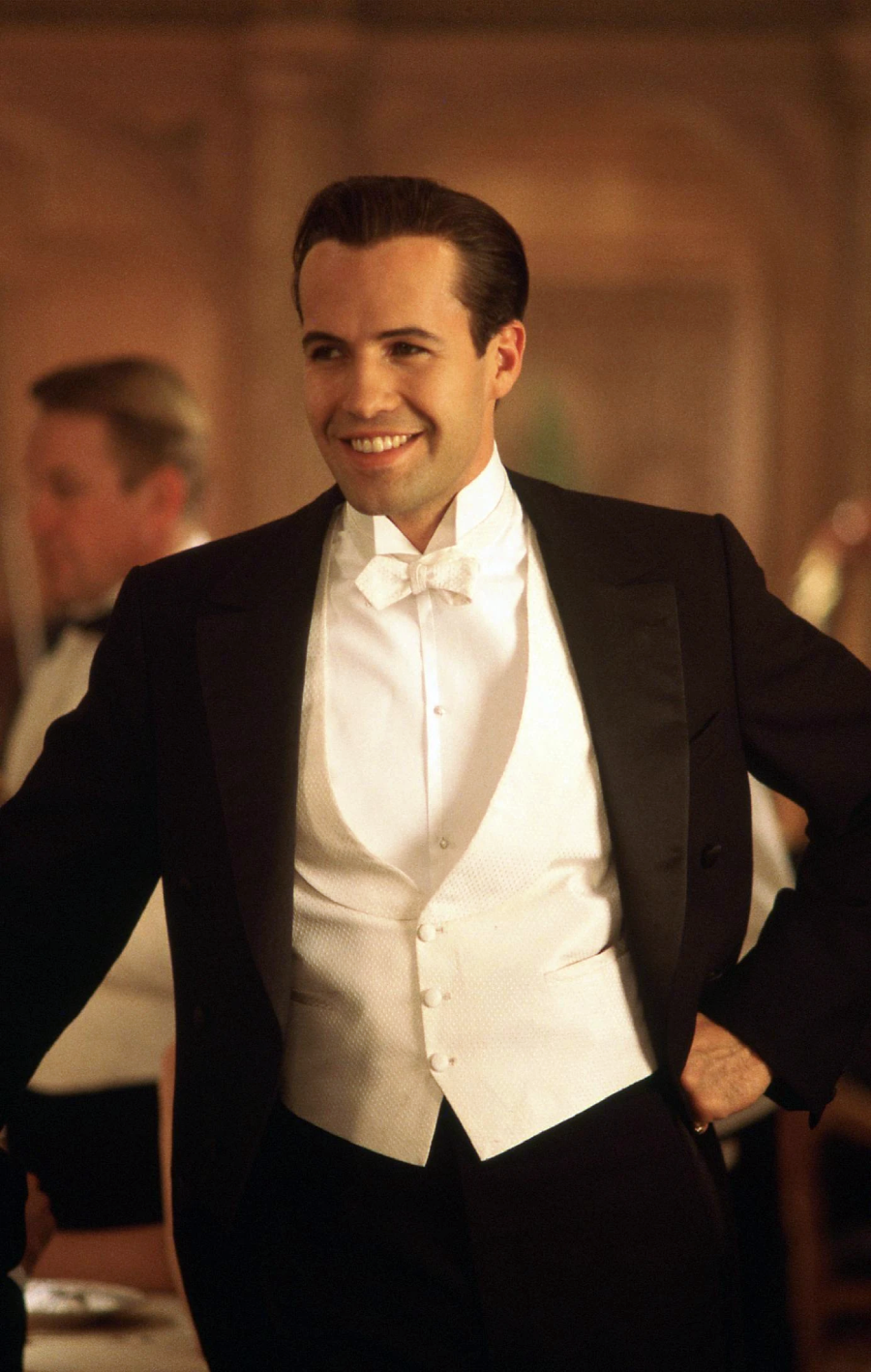 A man in formal evening wear, including a black tailcoat, white bow tie, and white waistcoat, smiles warmly at an indoor event with soft lighting and blurred people in the background.