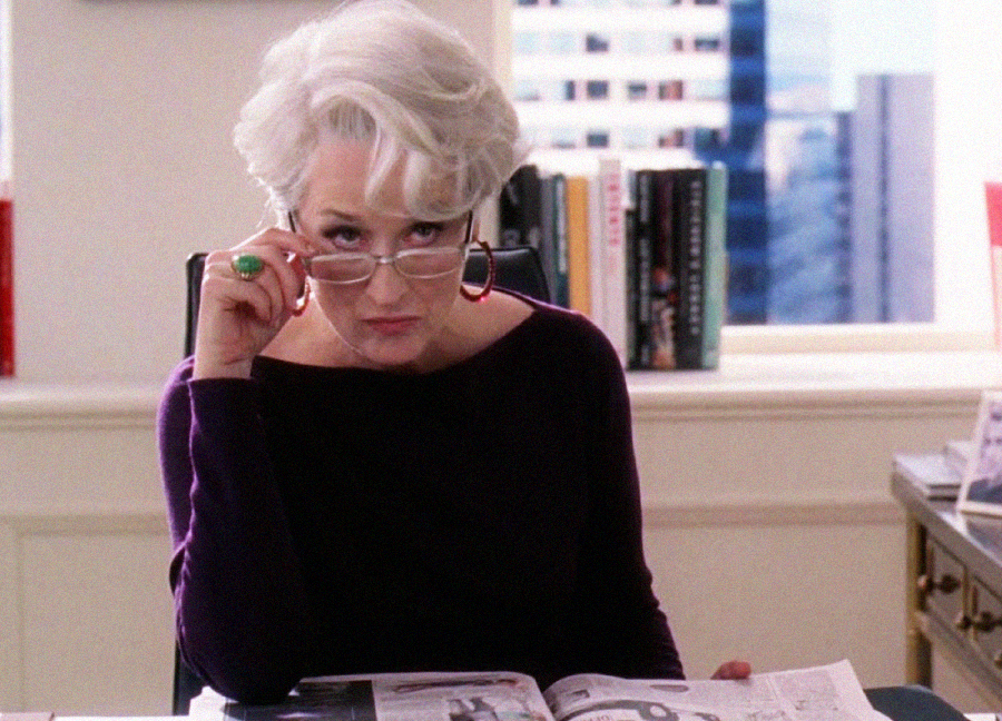 A woman with short white hair and glasses looks over the top of her frames with a stern expression, sitting at a desk with an open magazine in front of her, in a modern office with shelves and a window behind her.