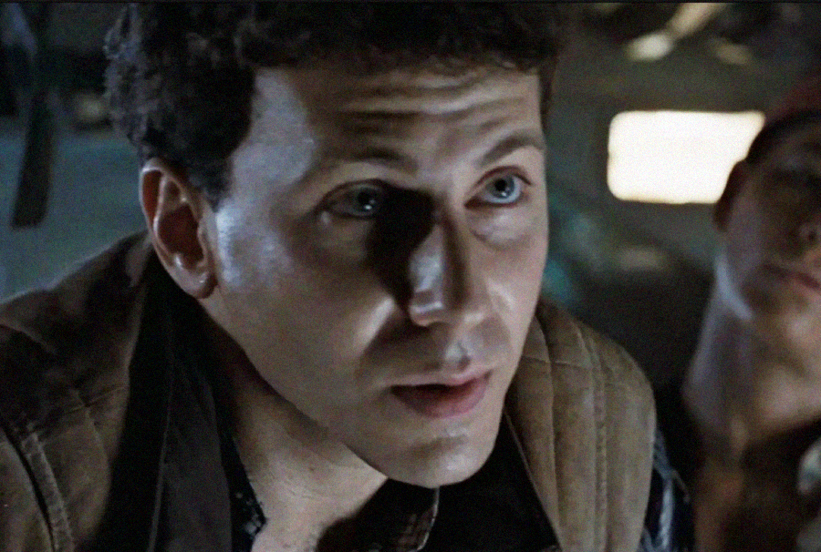 A close-up of a man with short dark curly hair and blue eyes, wearing a brown vest and looking slightly upwards with a serious expression. Another person is blurred in the background. The lighting is dim.