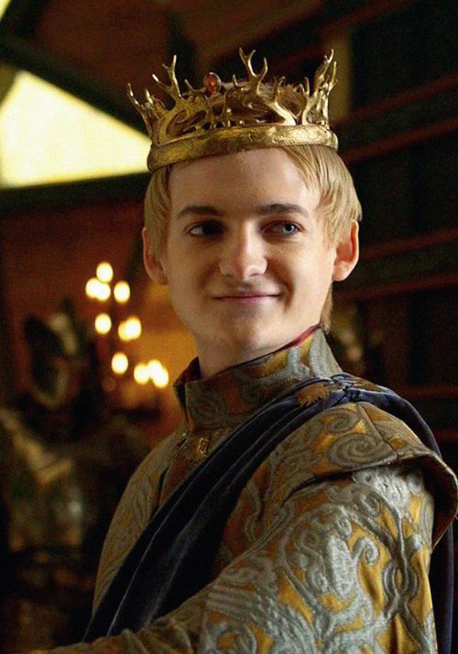A young man with short blond hair wears an ornate golden crown and richly embroidered medieval-style robes, standing in a dimly lit room with candelabras in the background. He is smiling slightly and looking to the side.