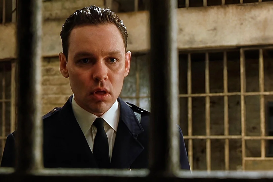 A man in a dark police uniform stands indoors, looking through metal prison bars with a serious, surprised expression. Blurred jail cell doors and stone walls are visible in the background.