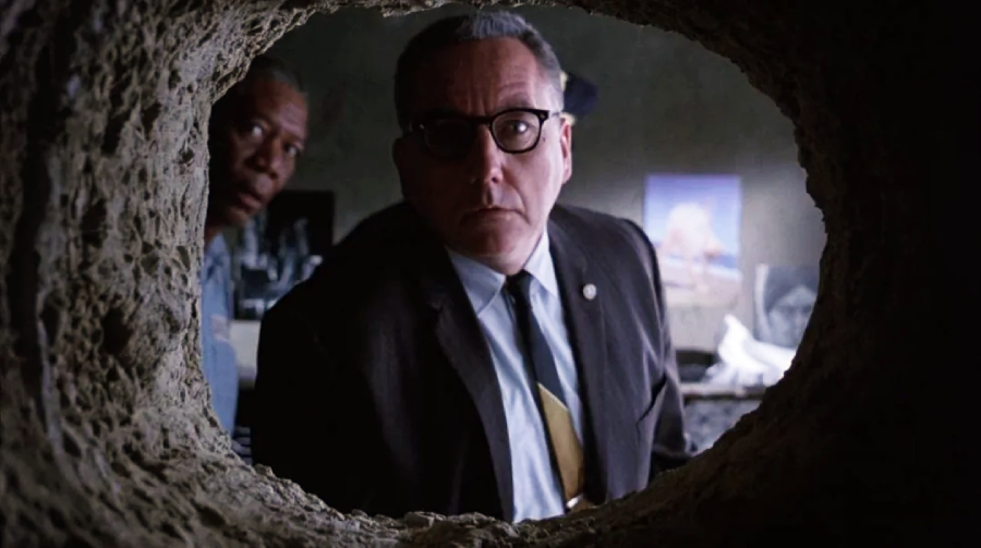 Two men peer into a rough hole in a wall from inside a dimly lit room, with posters and objects visible in the background. The man in front wears glasses and a suit, while the other stands behind him, partially visible.