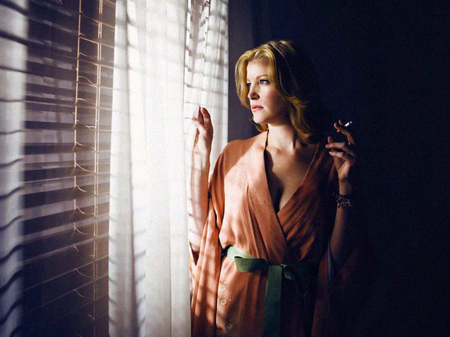 A woman in a peach robe with a green sash stands by a window with blinds, looking outside thoughtfully. Light filters through the blinds, creating shadows on the wall. She holds a cigarette in her right hand.