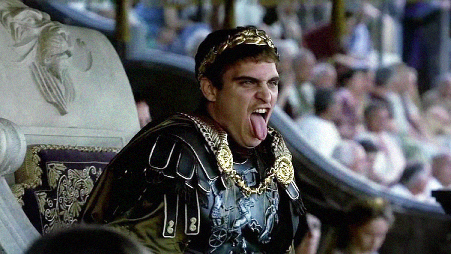 A man in ornate Roman-style armor and a gold laurel crown sits on a throne, sticking out his tongue with a playful or mocking expression. A blurred crowd is visible in the background.