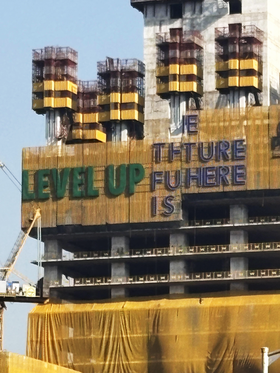 A high-rise building under construction features large green letters spelling "LEVEL UP" and a partially obscured sign reading "THE FUTURE IS HERE." Yellow scaffolding and cranes are visible around the structure.