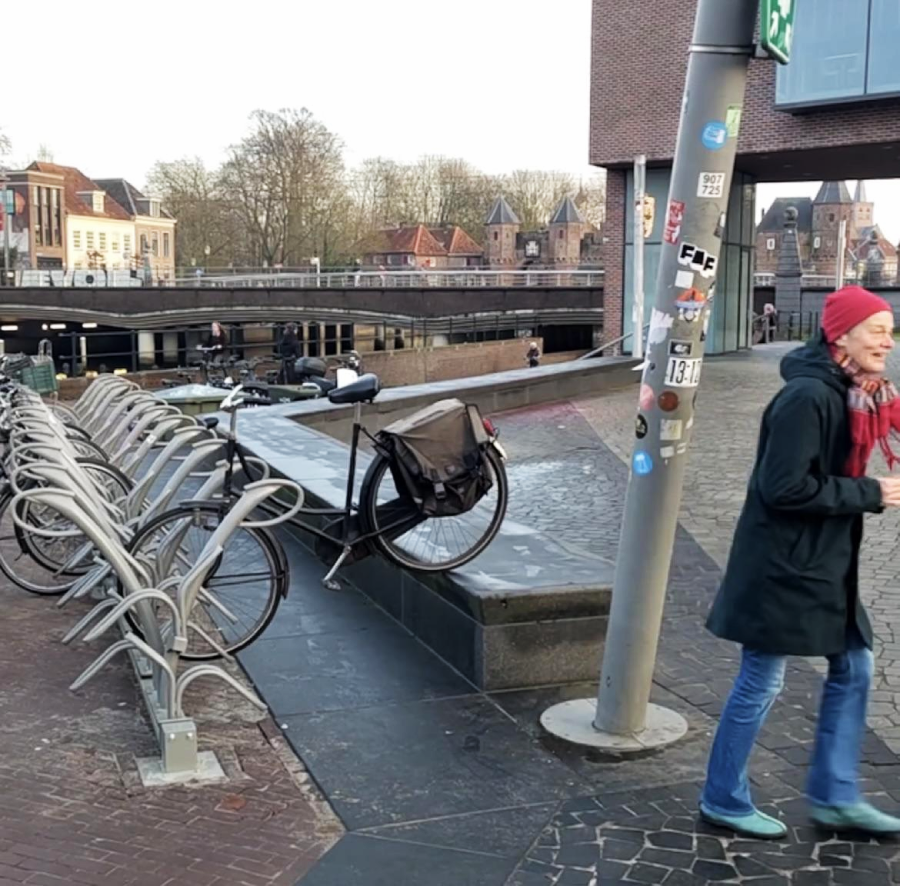 A woman in a red hat and blue coat walks past an empty bike rack, with one bicycle propped up on a raised concrete platform in an urban area with buildings and a bridge in the background.