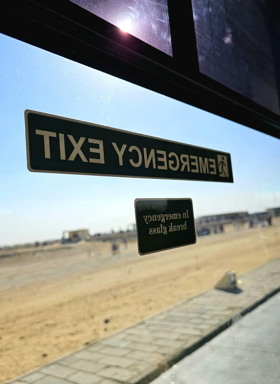 View from inside a vehicle looking out a window with stickers reading "EMERGENCY EXIT" and "In emergency break glass," showing a sandy, outdoor landscape and blue sky beyond.