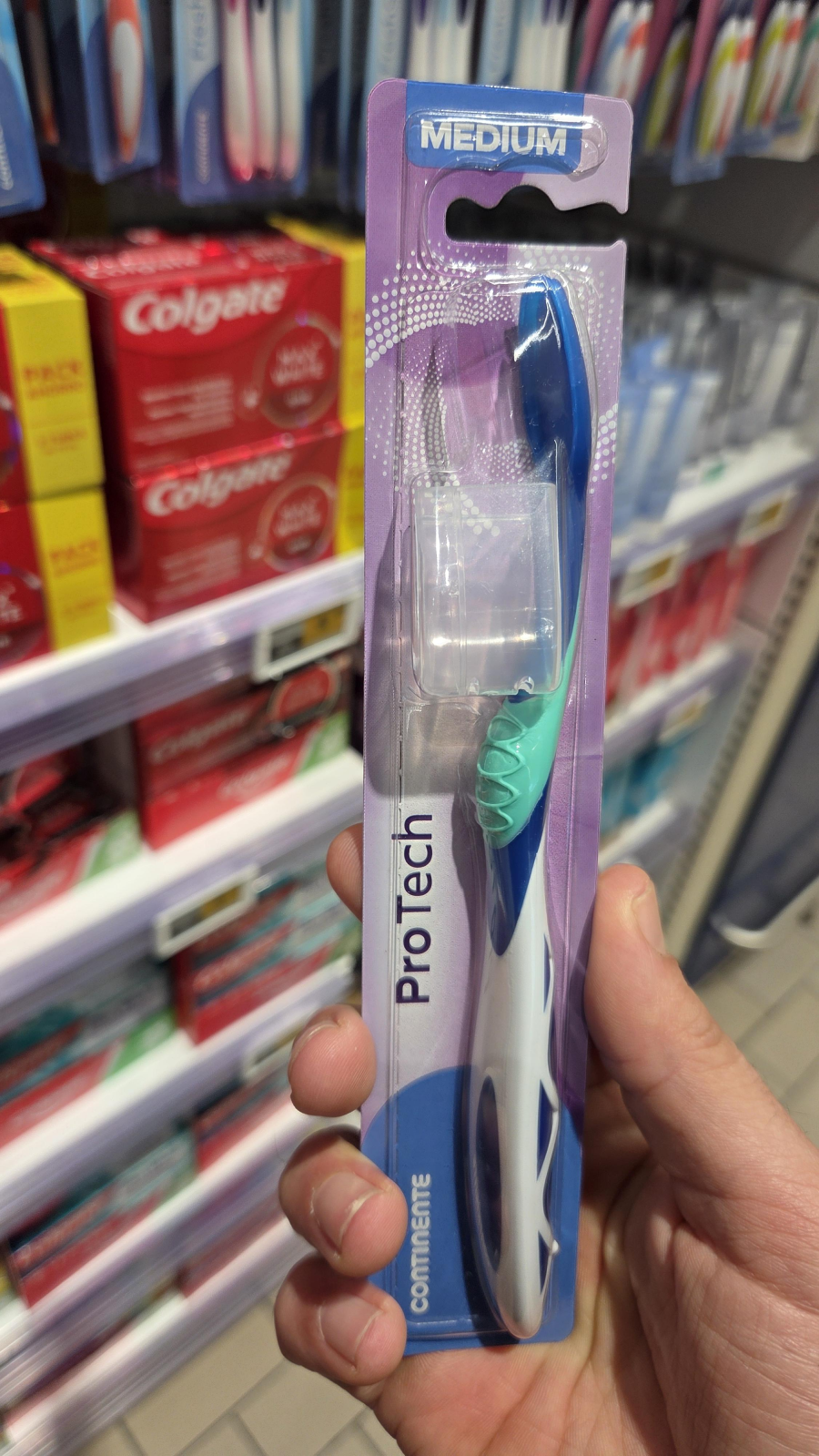 A hand is holding a packaged Pro Tech medium toothbrush in a store aisle, with shelves of Colgate toothpaste and other toothbrushes visible in the background.
