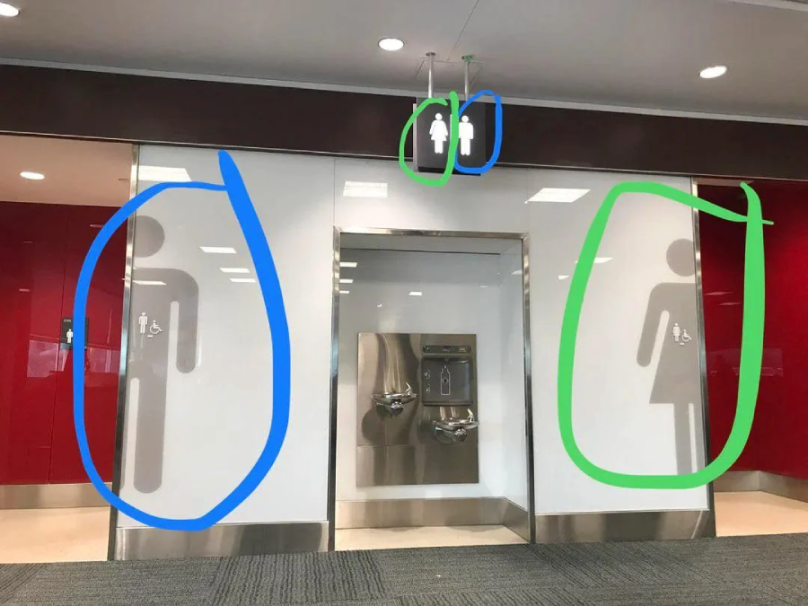 A restroom entrance with an oversized men's symbol on the left and an oversized women's symbol on the right, separated by drinking fountains; matching gender icons hang above.
