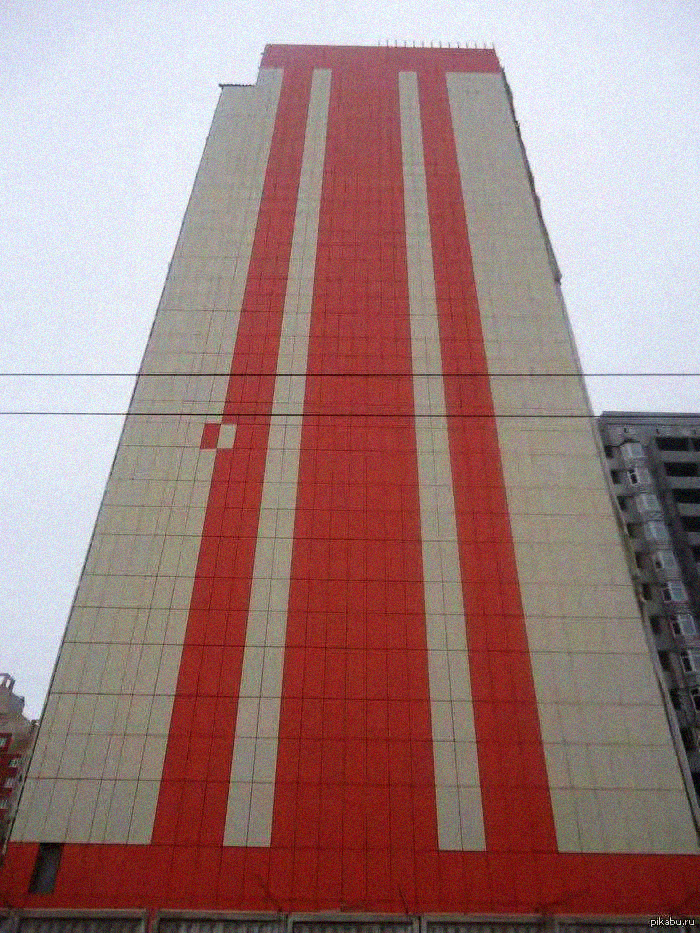 A tall building with a beige facade features bold vertical red stripes, including one wide central stripe and two narrower stripes on each side, viewed from the ground looking upward.