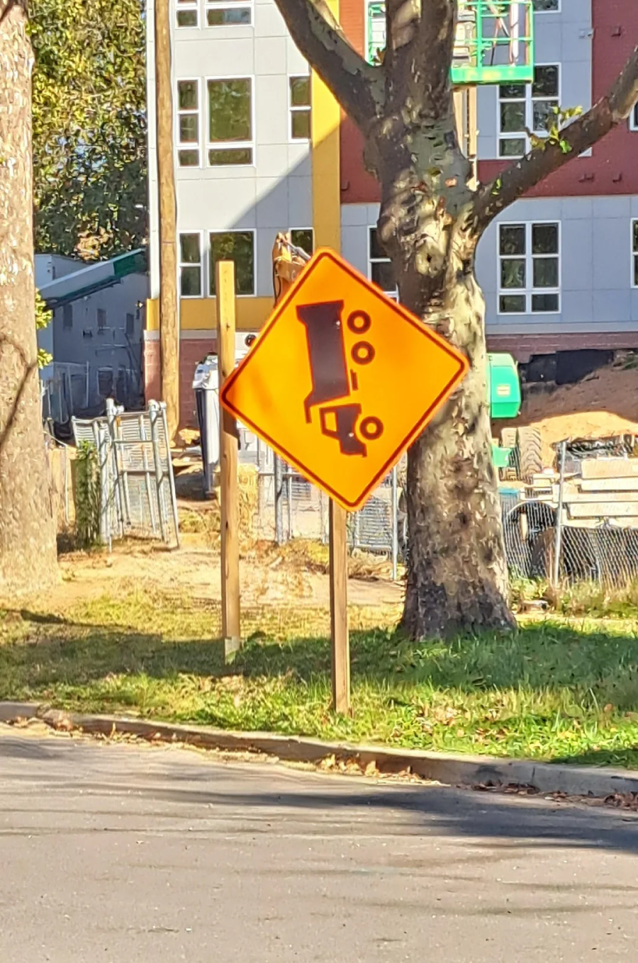 A bright orange road sign warns of trucks tipping over, positioned near a construction site with a tree, construction fencing, and a building in the background.