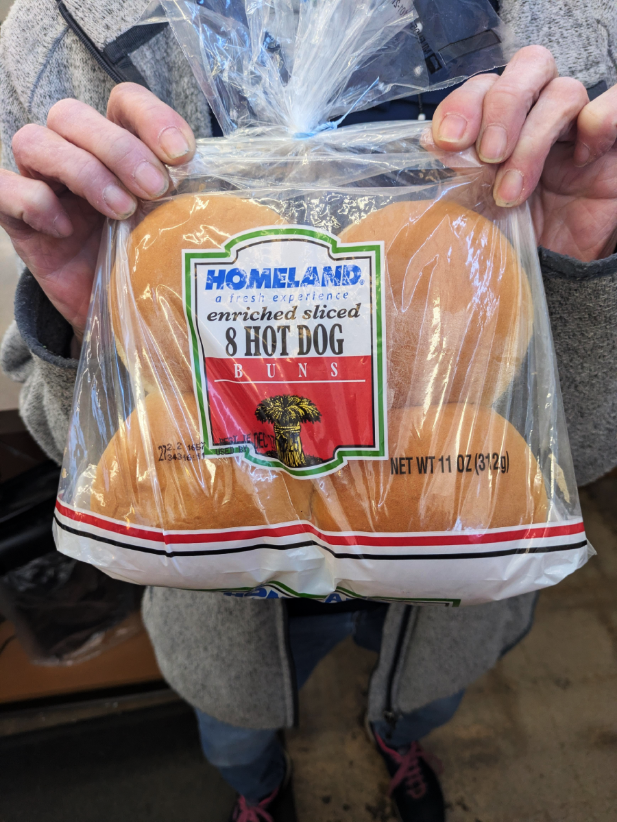 A person holds a package of Homeland brand enriched sliced hot dog buns, containing 8 buns in a clear plastic bag with red and white labeling.