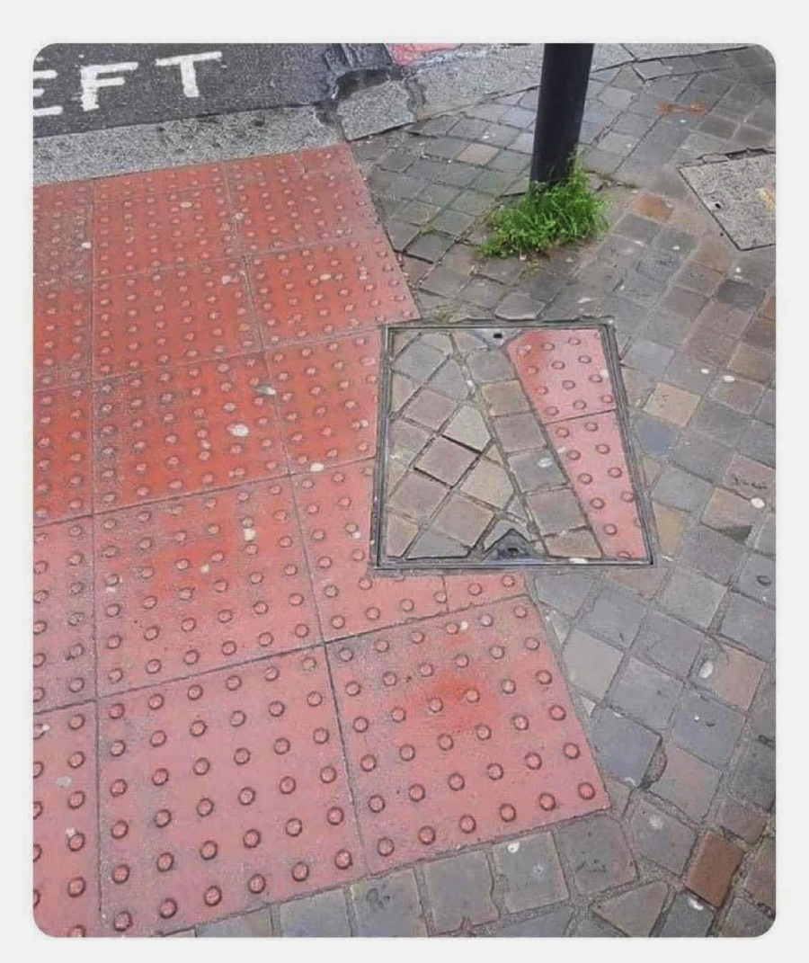 A sidewalk with red tactile paving meets a section of grey brick pavement; a utility cover is poorly placed, mixing both patterns and creating a jarring visual mismatch. Grass grows at the base of a black pole nearby.