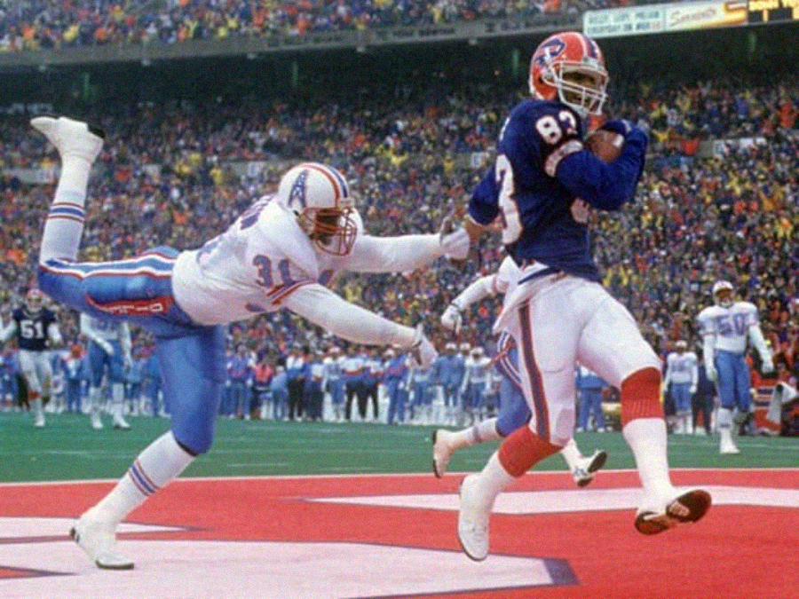 A football player in a blue Buffalo Bills uniform runs into the end zone with the ball as a Houston Oilers defender dives and stretches out, trying to tackle him, during a game in a crowded stadium.