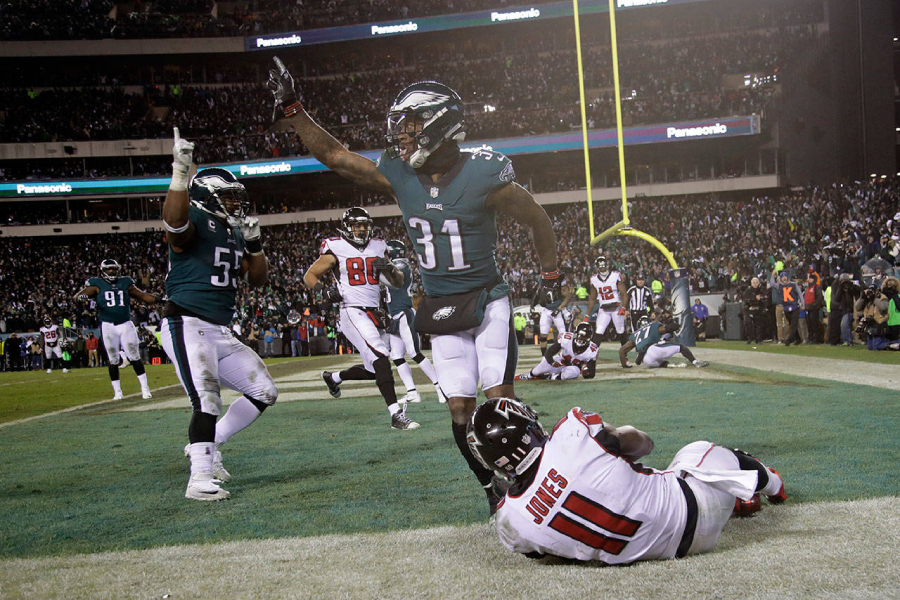 Philadelphia Eagles football players celebrate in the end zone as an Atlanta Falcons player sits on the ground during a night game, with a cheering crowd and stadium lights in the background.
