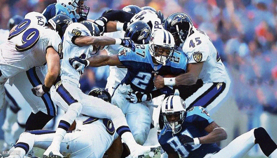 A football player in a blue and white uniform runs with the ball as several defenders in white and purple uniforms try to tackle him during a game, creating a dynamic and intense action scene.