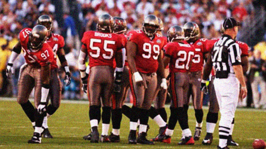 A group of football players in red and brown uniforms huddle on a field near a referee in a black-and-white striped shirt; a crowd fills the stands in the background.
