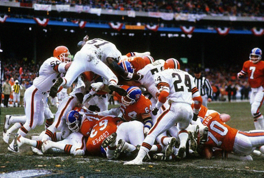 A chaotic football scene shows players in orange and white uniforms piling up at the goal line during an intense game, with one player leaping over the top as others scramble for the ball. A packed stadium is visible in the background.