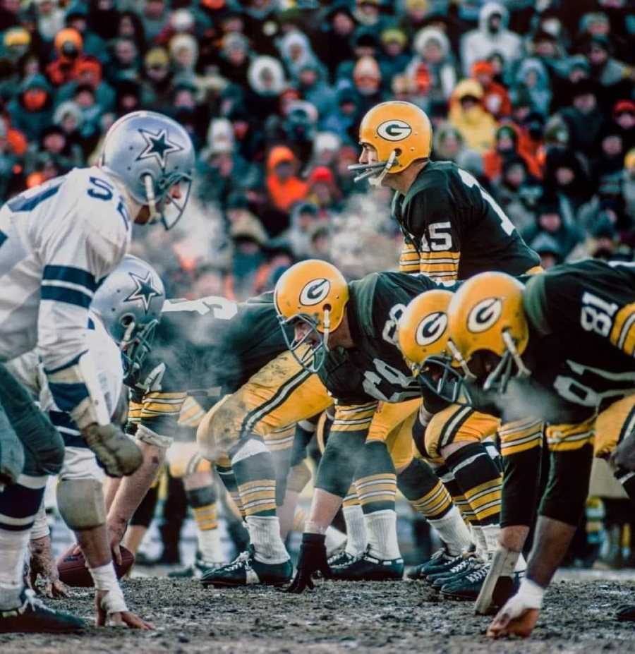 Two American football teams, the Green Bay Packers and Dallas Cowboys, face off on a cold day, visible breath in the air, as players line up at the line of scrimmage, with bundled fans watching in the stands.