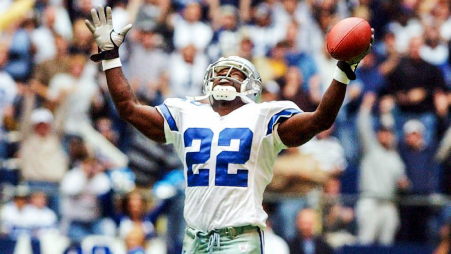 A football player in a white and blue Dallas Cowboys uniform, wearing the number 22, raises his arms in celebration while holding a football. The background shows a cheering crowd in a stadium.